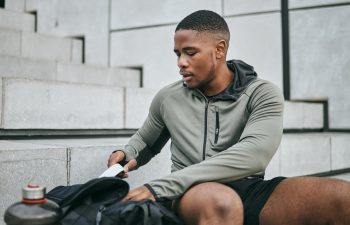Man in a gray hoodie sitting on concrete steps, organizing his backpack with a white towel next to him. A water bottle is visible in the foreground.