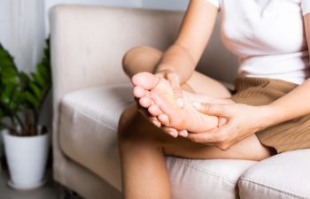Foot pain, Asian woman sitting on sofa feeling pain in her foot at home, female suffering from feet ache use hand massage relax muscle from soles in home interior, Healthcare and podiatry medical